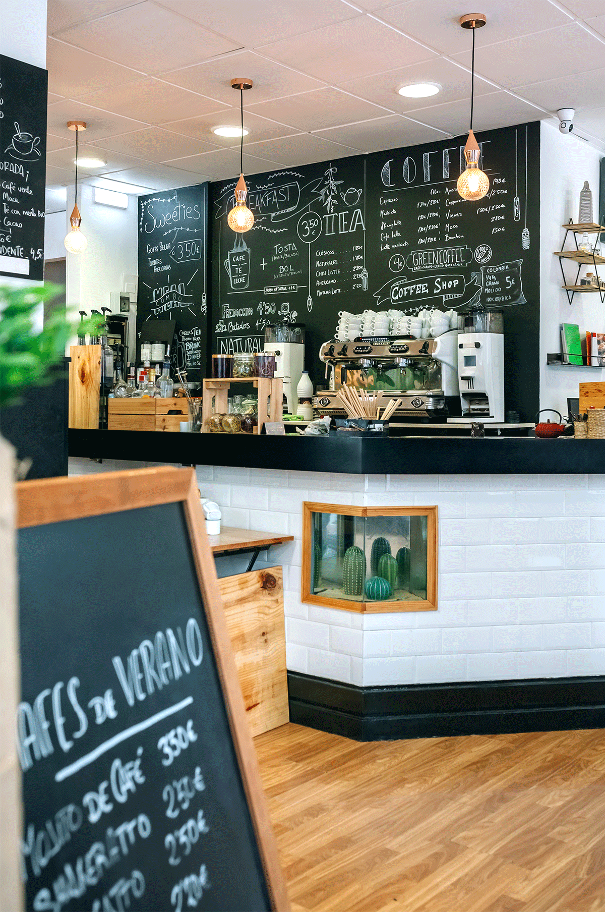 Kafébar med svart tavlemeny, espresso-maskin, kopper og hengende lyspærer i moderne interiør.