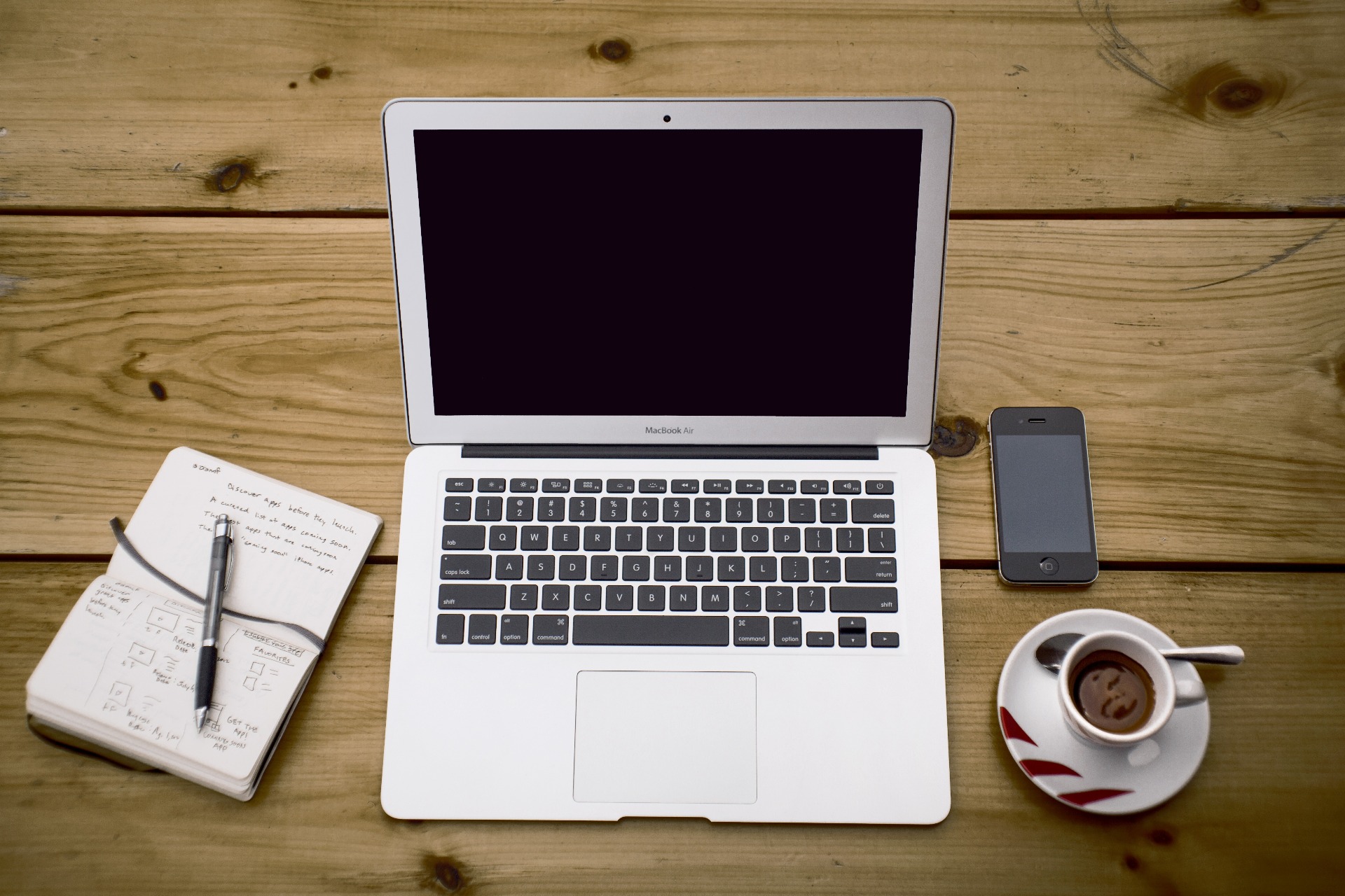 Fehér MacBook Air laptop nyitott képernyővel, jegyzetfüzettel, tollal és kávéscsészével faasztalon.