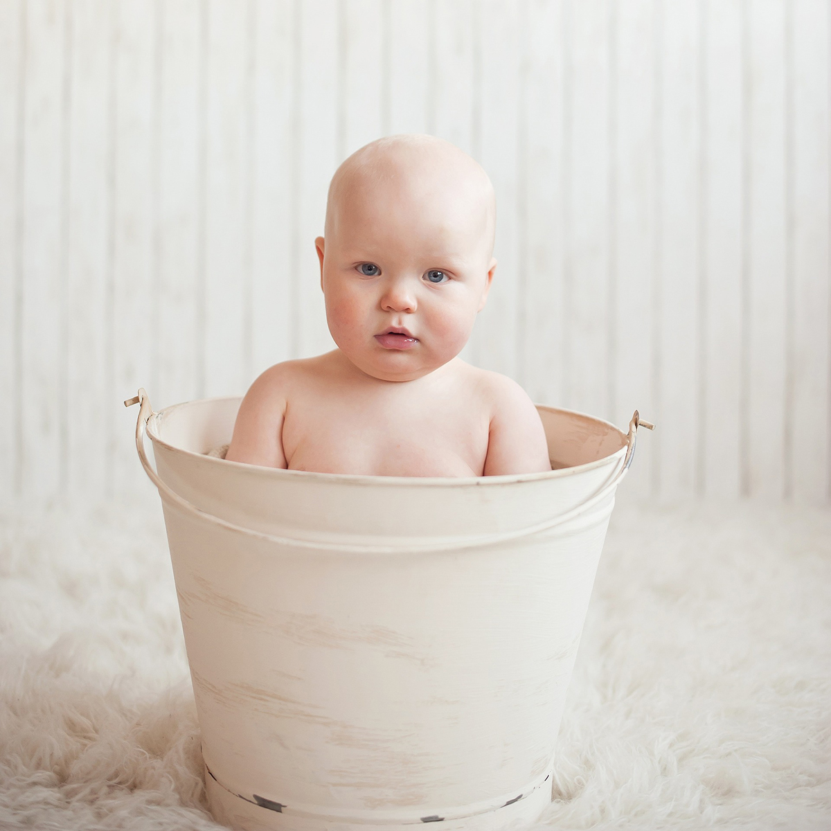 Bare baby with blue eyes sitting in a white bucket on soft fur, neutral studio background.