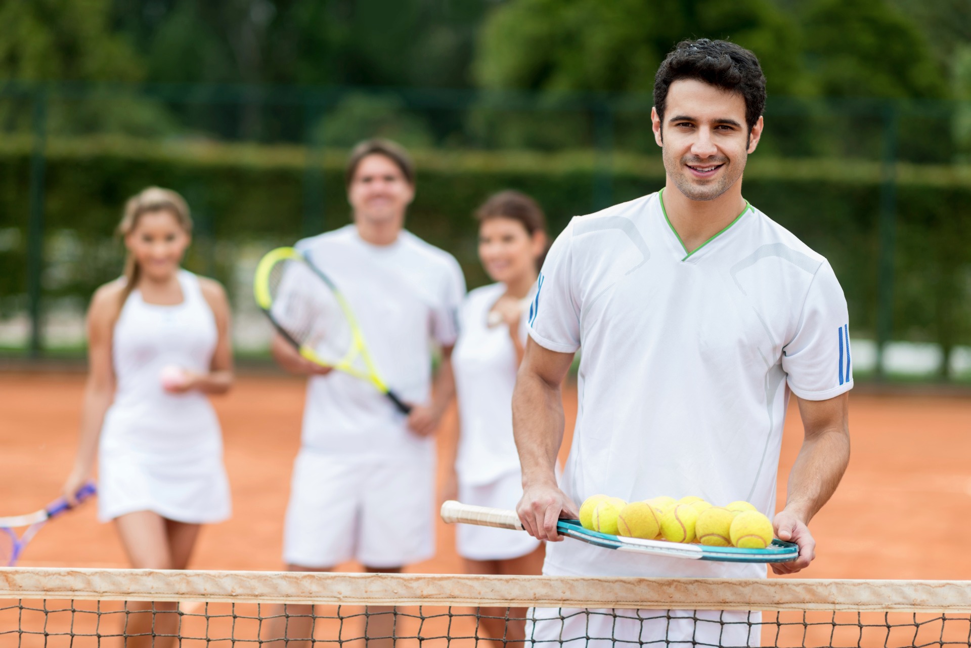 Hombre sonriente con raqueta y pelotas de tenis, mientras tres personas desenfocadas juegan en la pista.