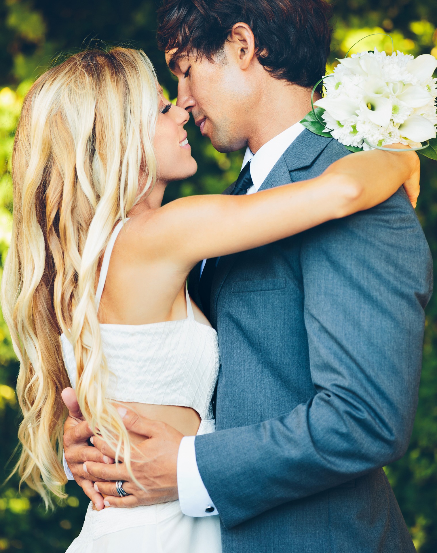 Pareja abrazada al aire libre, él con traje azul y ramo blanco, miradas cercanas y sonrisas.